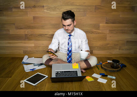 A barefoot businessman sits on the floor against a wall with cup of coffee and smartphone. Using laptop and gadgets. Calm young man in funny clothes Stock Photo