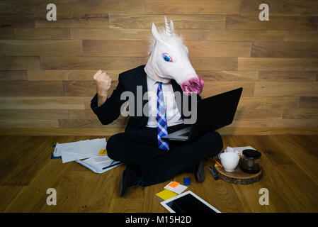 Young man in funny horse mask sits on the floor with cup of coffee and works with laptop. Angry and dissatisfied unicorn in a suit shows fist Stock Photo