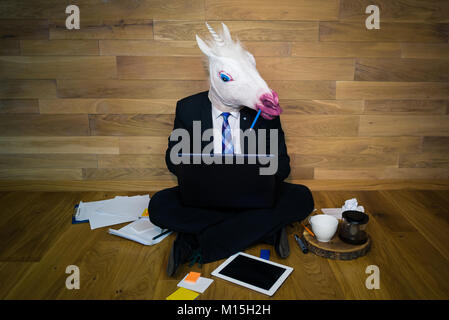 Young man in funny horse mask sits on the floor and works with laptop.  Serious unicorn in a suit enthusiastically works with a pencil in the teeth Stock Photo