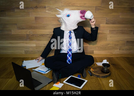 Young man in mask drinking a coffee. Unicorn in a suit and tie working with documents and using laptop and gadgets on wooden background Stock Photo