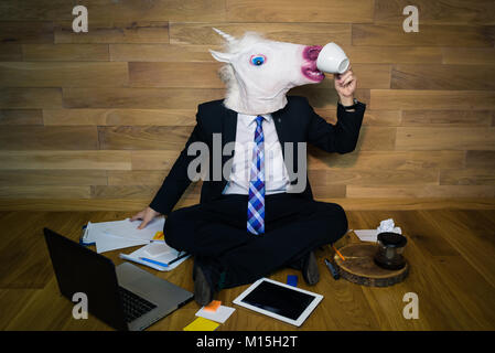Young man in mask sits on the floor against a wall and drinking coffee. Unicorn in a suit works with documents on wooden background Stock Photo