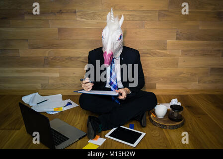 Young man wearing rubber mask sitting on the floor and works with the documents. Unicorn in a suit using laptop and gadgets Stock Photo