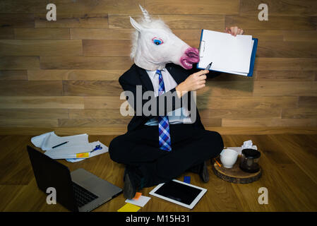 Unicorn in a suit shows a white empty sheet with copy space. Any text possible. Young man in mask sits on the andc works with laptop Stock Photo