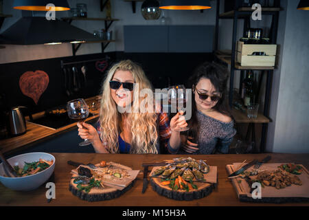 Two funny girlfriends holds glasses of wine at the bar counter. Happy women sits at the table with food. Young people enjoying time together Stock Photo