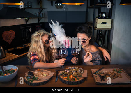 Three young people has dinner together and holds glasses of wine. Unicorn with two beautiful women. Crazy evening in modern home kitchen Stock Photo