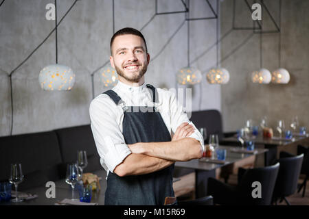 A young, smiling waiter in a restaurant, standing next to the tables with a glass of wine. He wears an apron, looks confidently, folded his arms over  Stock Photo