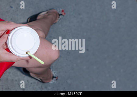 a woman's hand holds a paper cup of coffee. from the cup, stick out the straw. the girl is sitting in a red dress. a paving stone. beautiful woman's l Stock Photo