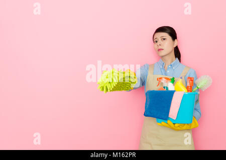 seriously attractive housewife using feather duster tool pointing camera and holding many cleaning equipment standing in pink background. Stock Photo