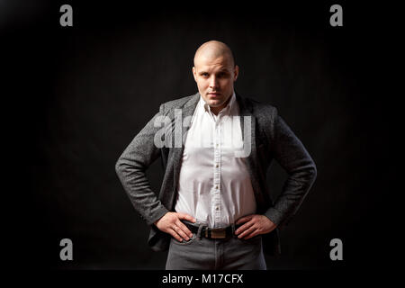 A  bald man businessman  in a white shirt, gray suit stands,  looks seriously and holds his hands at the waist on a black isolated background Stock Photo