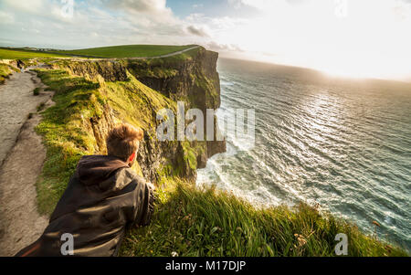 Young man laying down on the edge of the cliffs on the Coastal footpath walk, Cliffs of Moher, huge sea cliffs located at the southwestern edge of the Stock Photo