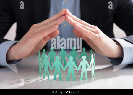 Close-up Of A Businessperson's Hand Protecting Blue Paper Cut Out Figures On White Background Stock Photo
