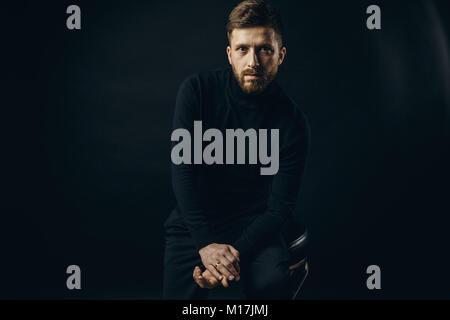 Bearded man wearing black looking unemotionally at camera on black background of studio. Stock Photo