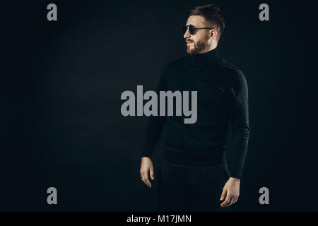 Bearded man in elegant black turtleneck and flip sunglasses standing with confidence on black. Stock Photo