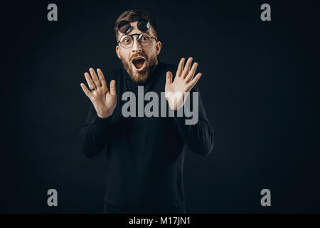 Young bearded man in black looking with surprise at camera wearing round flip-up glasses on black. Stock Photo