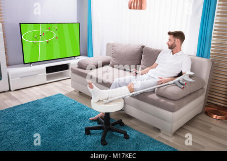 Young Man With Broken Leg Sitting On Sofa Watching Football Match On Television Stock Photo