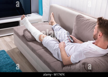 Young Man With Broken Leg Lying On Sofa Holding Remote Near Television Stock Photo