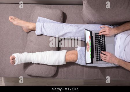 High Angle View Of A Man With Plastered Leg Video Conferencing With Doctor On Laptop Stock Photo