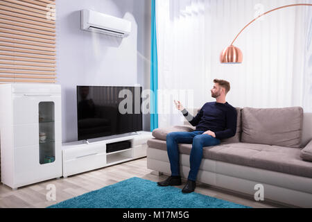 Young Man Sitting On Sofa Using Air Conditioner At Home Stock Photo