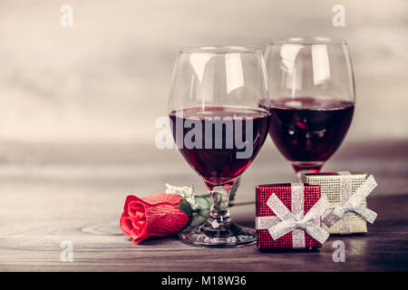 Two glasses of red wine, rose and gift with decoration by red hearts on wooden table. Valentines day celebration concept. Copy space. Stock Photo