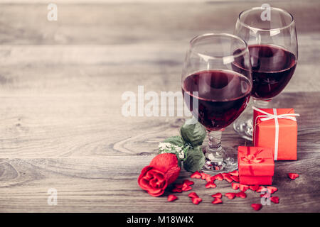 Two glasses of red wine, rose and gift with decoration by red hearts on wooden table. Valentines day celebration concept. Copy space. Stock Photo