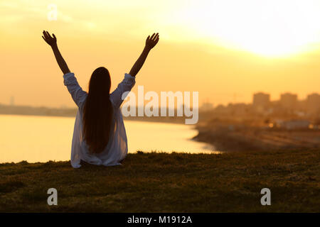 Back view backlight portrait of a happy single woman seeing the city at sunset and raising arms with a warm light in the background Stock Photo