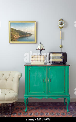 Interior shot of green vintage sideboard with old radio, stack of old books, and table lamp on background of off white wall with hanged painting Stock Photo
