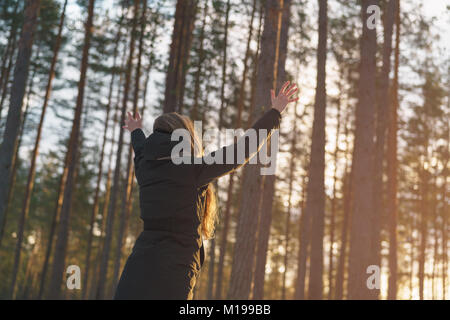 teen girl raised hands from behind in winter pine forest in sunset Stock Photo