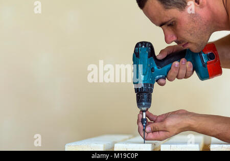 Hand of a worker screws a screw in a wooden board with a cordless screwdriver. Man carpenter at handmade work Stock Photo