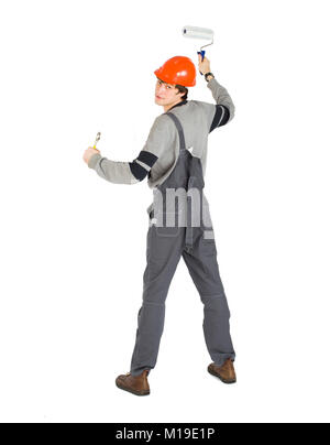 A young man in working grey clothes and orange hard helmet painting the walls in the room. Stock Photo
