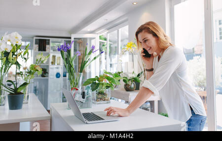 Female florist standing at her flower shop counter using mobile phone and laptop to take orders for her store. Woman flower shop owner taking online o Stock Photo
