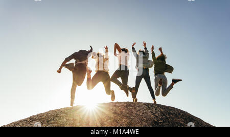 Rear view of group of happy friends having fun on mountain top. Men and women jumping on mountain top against sunset. Stock Photo