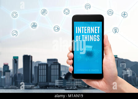 Hand holding mobile with Internet of things (iot) word on screen with icon feature glowing in air with blur city building background,Digital Lifestyle Stock Photo