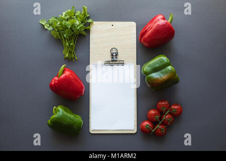 top view of various fresh peppers, tomatoes, parsley and blank paper sheet with cutting board on grey Stock Photo