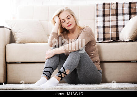 Depression. Middle aged barefoot woman sitting at the floor embracing her knees, near sofa at home, her head down, bored, troubled with domestic viole Stock Photo