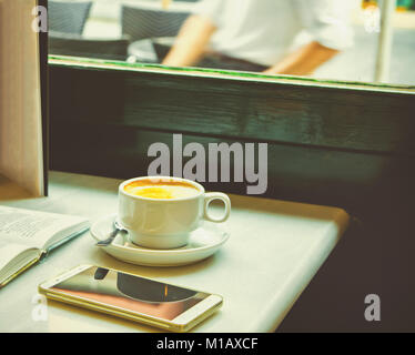 White Cup with Cappuccino on Table with Open Book Smartphone on Cafe Table by Window. Wooden Chair Cozy Atmosphere. European Style. Lifestyle Photo To