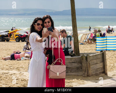 Two smartly dressed young millennial women taking selfies together using smartphones on a beach in late summer. Bournemouth Dorset England UK Britain