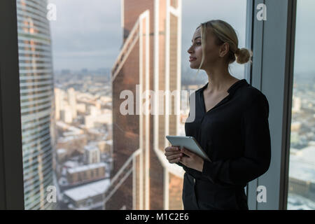 Business woman holding a tablet and standing in a modern office. Panoramic windows background. Stock Photo