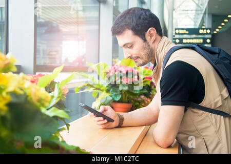 young man wearing t-shirt and travel vest with backpack on his back waiting for his plane in departure lounge using his smartphone at window with flow Stock Photo