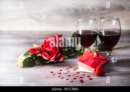 Two glasses of red wine, roseand gift with decoration by red hearts on wooden table. Valentines day celebration concept. Copy space. Stock Photo