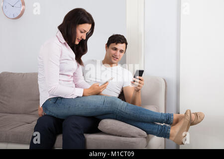 Young Couple Sitting On Sofa Using Mobile Phone At Home Stock Photo