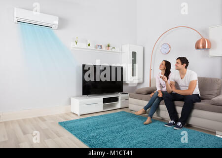 Smiling Young Couple Sitting On Sofa Using Air Conditioner Stock Photo