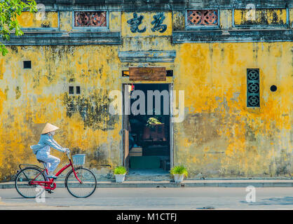 Vietnamese lady everyday life  riding a bicycle in front of an old traditional building in hoi an town. Vietnam. Stock Photo