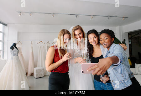 Woman in wedding gown with female friends taking selfie in bridal boutique. Beautiful bride in elegant wedding dress and friends making self portrait  Stock Photo