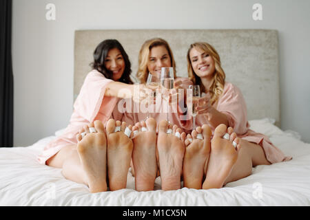 Female friends celebrate a bachelorette party of bride. Three female sitting on bed and toasting champagne glasses at home. Focus on legs with female. Stock Photo