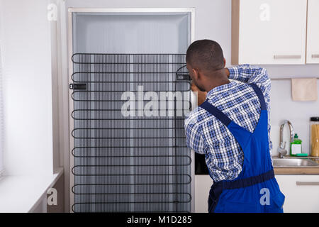 Serviceman In Overall Working On Fridge With Screwdriver In Kitchen Stock Photo