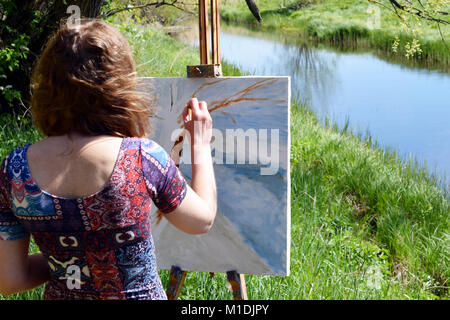 Rear view of artist paints a beautiful summer landscape with river. Painting on canvas is not ready. Stock Photo