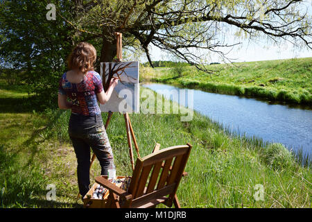 Woman artist paints landscape painting of small river. Painter rear view. Stock Photo