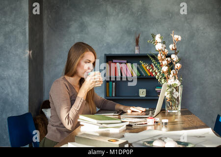 Young  Woman Sitting at Office, Drinking Coffee and Using Laptop Stock Photo