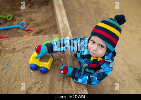 Very serious child playing with toys and have fun in the sandbox. Beautiful kid plays on the playground with small car and looking in camera. Stock Photo