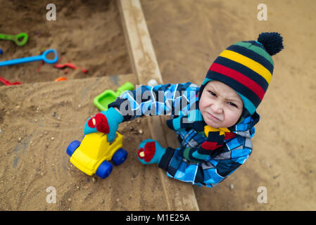 Very serious child playing with toys and have fun in the sandbox. Beautiful kid plays on the playground with small car and looking in camera. Stock Photo